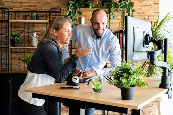 Frau und Mann besprechen gemeinsam Inhalte an einem höhenverstellbaren Schreibtisch mit Monitor im Büro.